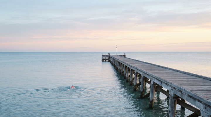 Timber jetty into a calm sea at sunrise with a lone swimmer